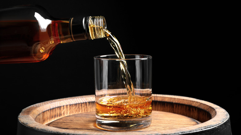 Bourbon being poured from a bottle into a glass sitting atop a wooden barrel