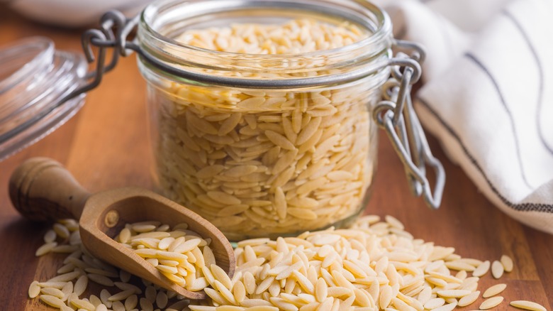 Orzo pasta in a jar with some spilled on the table with a wooden scoop