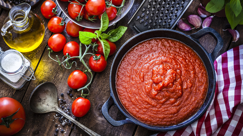 Marinara sauce in a pot with oil, herbs, and tomatoes on the side