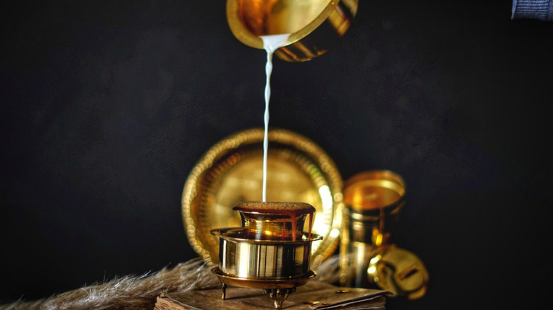 Pouring milk from a gold bowl into a cup of freshly brewed kaapi