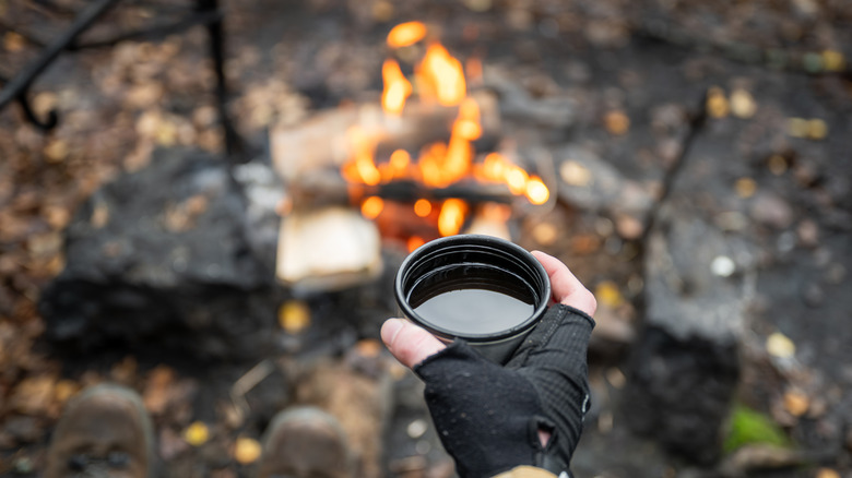 A hand holding a cup of coffee in front of a campfire