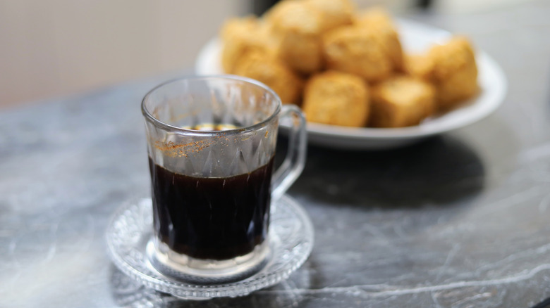 A small glass of black Kopi Tubruk with a plate of snacks in the background