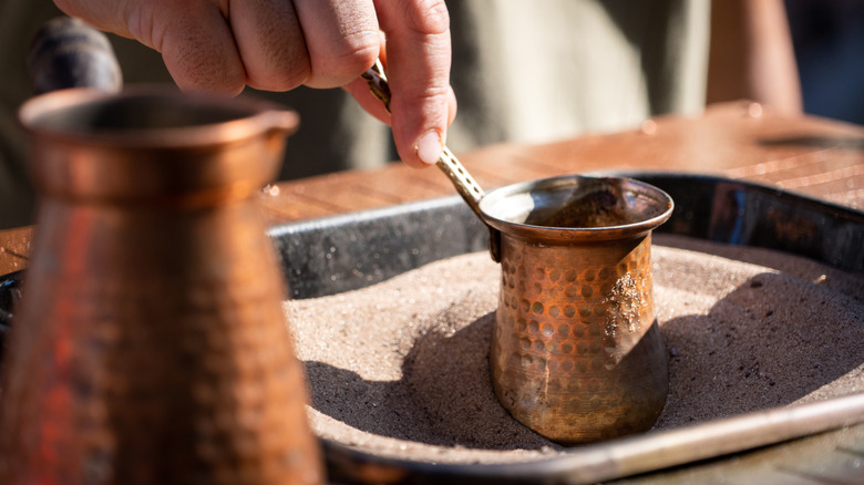 A hand holds the handle of a cezve and swirls it in hot sand