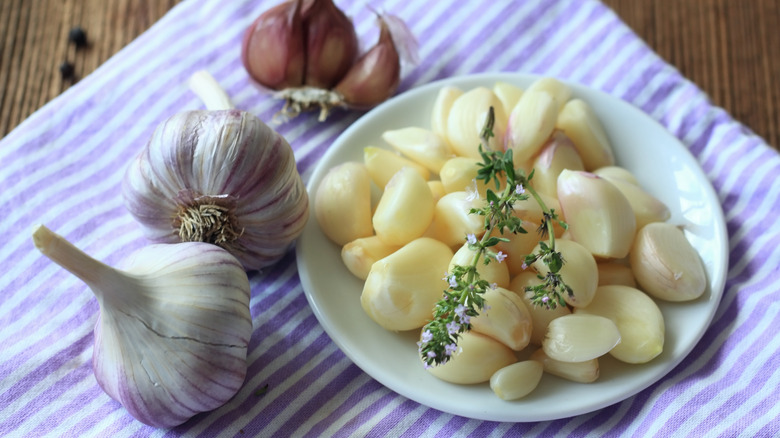 A dish of peeled garlic cloves with a thyme sprig on top over a striped tea towel, surrounded by whole cloves of garlic