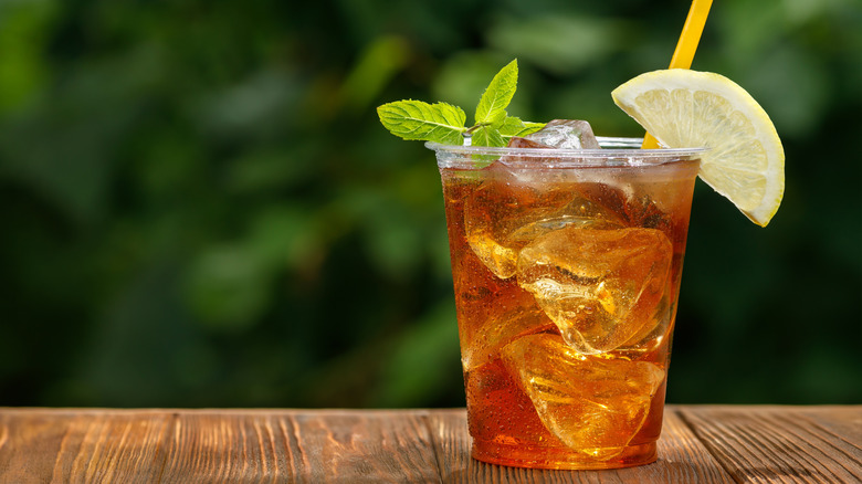 A plastic cup filled with iced tea, topped with a lemon and a sprig of mint, sitting on wooden table