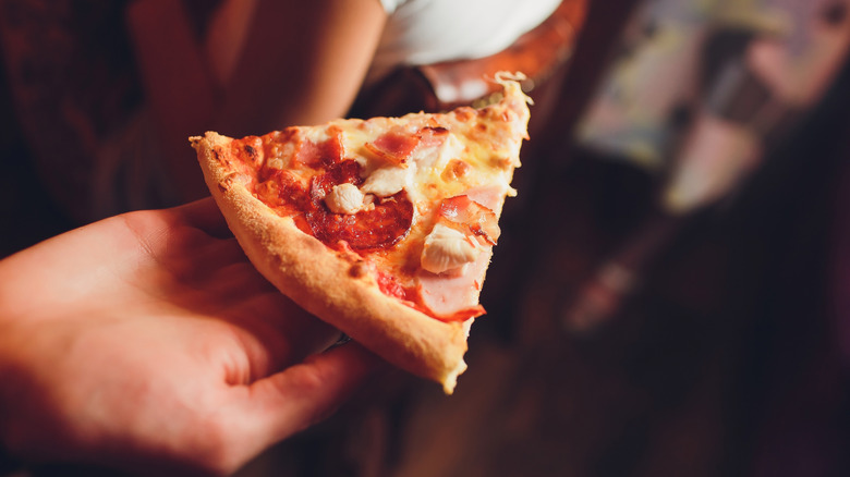 Caucasian hand holding slice of pizza with cheese and meat