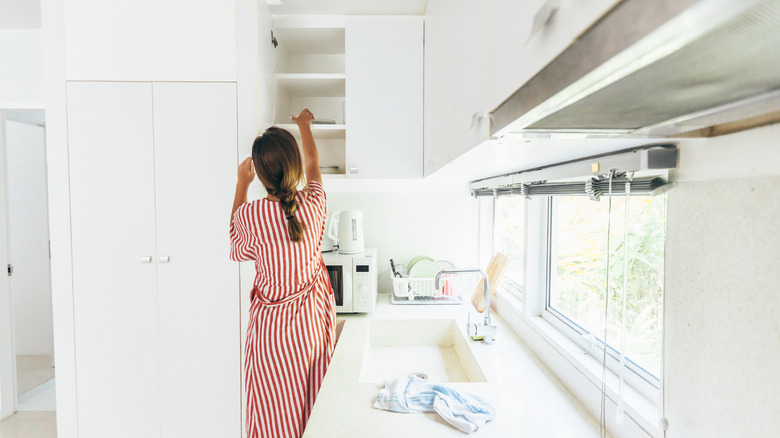 A woman cleans the shelves in a white, naturally lit kitchen