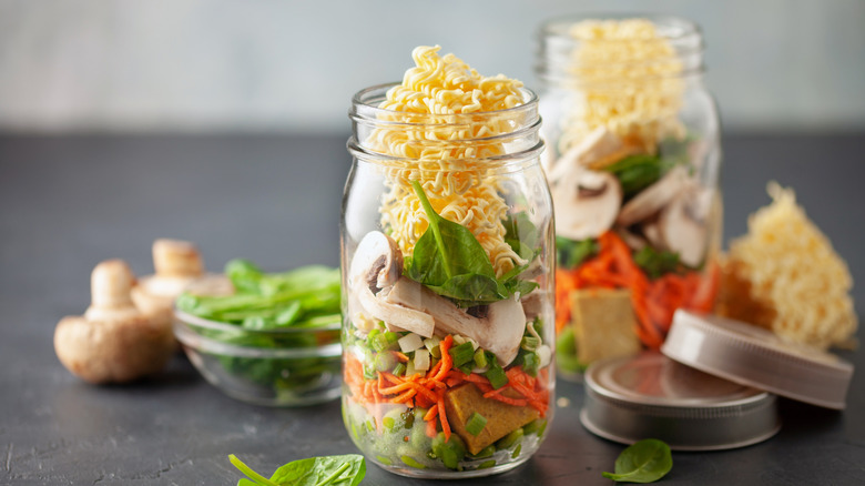 Two Mason jars are packed with soup ingredients like ramen, mushrooms, shredded carrots, and spinach.