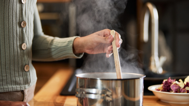 A person's hand is seen stirring a pot of steaming soup on a stove with a wooden spoon.