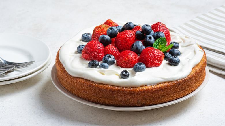 A sponge caked topped with whipped cream and berries on a table surrounded by plates with fork and striped napkins behind.