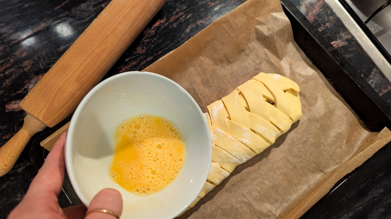 An individual holding a bowl of egg wash over pastry dough with rolling pin nearby.