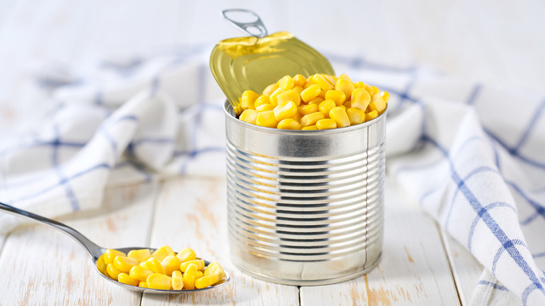An open can of corn with a spoon and tablecloth next to it