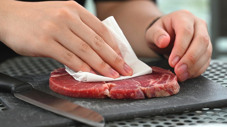 Hands drying a raw steak with a folded paper towel on a plastic cutting board