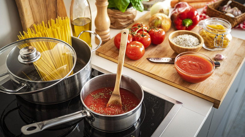 A busy kitchen tableau with sauce and pasta on the stove with other dinner ingredients on a cutting board nearby.