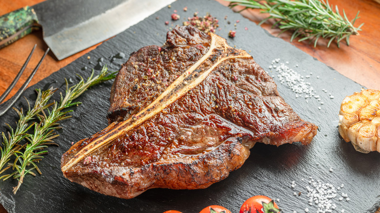 A seared t-bone steak on a slate board surrounded by herbs and salt.