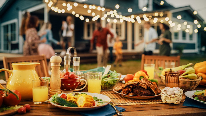 Outdoor dining table with various foods at backyard barbecue with people in background.