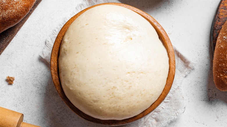 A bowl of raw bread dough surrounded by baked loaves of bread, a rolling pin, and a bowl of flour