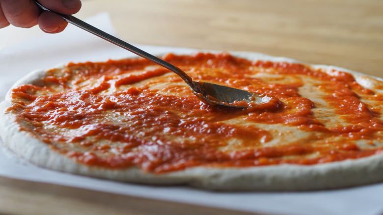 A hand spreading fresh, red pizza sauce on fresh pizza dough with a spoon.