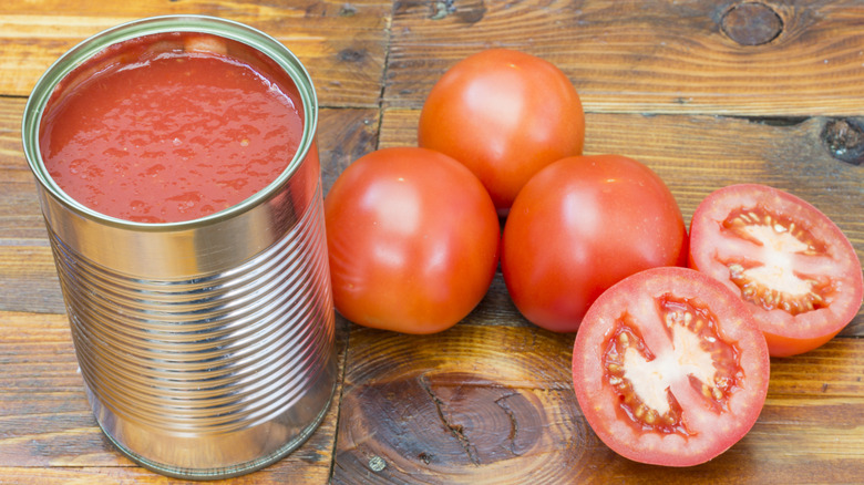 An open, unlabeled can of crushed tomatoes on a rustic wooden table with fresh tomatoes, one sliced in half, next to it.