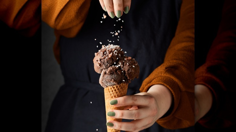 Person with painted nails sprinkling salt on a chocolate scoop of ice cream in a cone