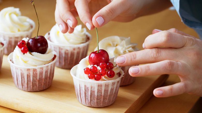 A person's hands are seen adding fruit to cupcakes.