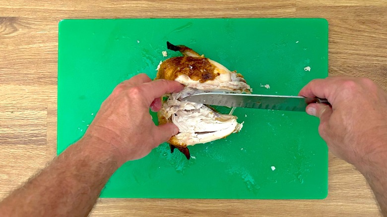 Carving the breast off a rotisserie chicken with a knife that is resting on a cutting board