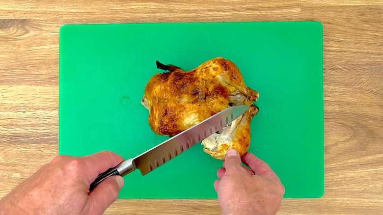 Removing the thigh and leg from a rotisserie chicken with a knife while it is resting on a cutting board