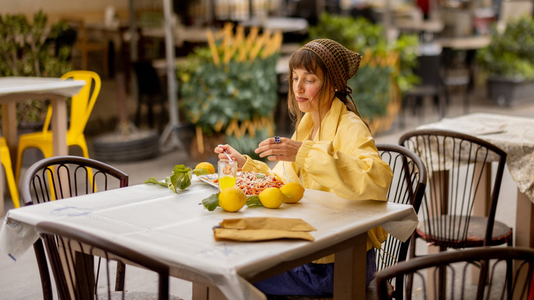A woman in a yellow jacket enjoys a plate of pasta alone at a street cafe under large umbrellas