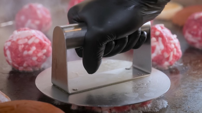 Cook uses a metal burger press to flatten raw ground beef balls on a hot griddle