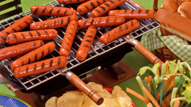 A hibachi grill with hot dogs surrounded by potato chips, vegetables, and hot dog buns