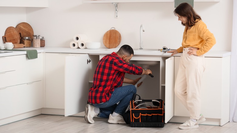 two people standing by kitchen sink while man works underneath it