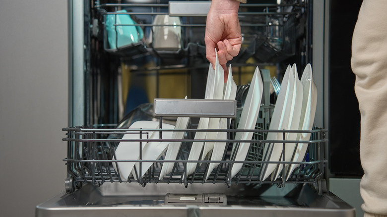 A hand reaches into a dishwasher to unload white plates