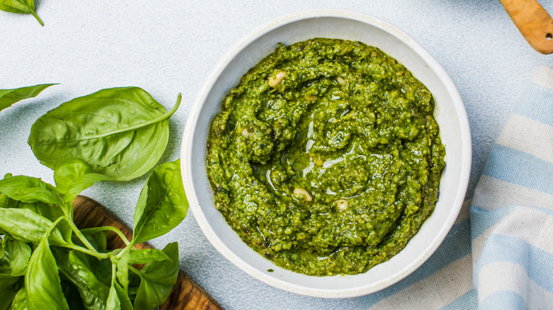 Pesto in a white bowl next to basil leaves on a cutting board