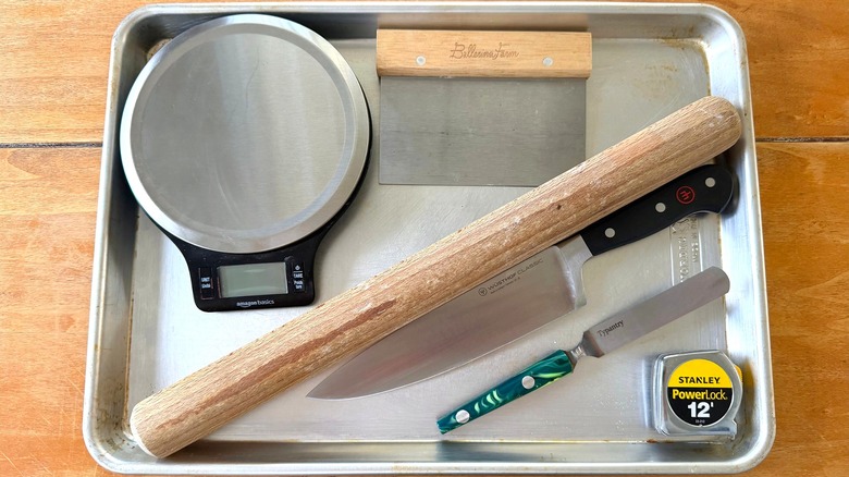 a sheet tray with a scale, bench scraper, rolling pin, knife, offset spatula, and tape measure