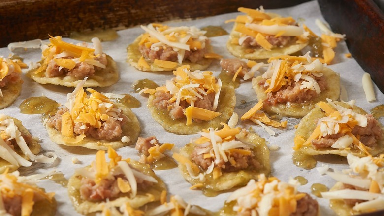 Individual round tortilla chips covered in refried beans, shredded cheese, and green salsa on a parchment-lined sheet pan