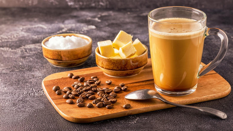 Keto coffee in glass atop wooden cutting board next to small wood bowls of coconut oil and cubes of butter.
