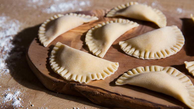 Freshly made ravioli sit on a rustic wooden board on a flour-sprinkled table.