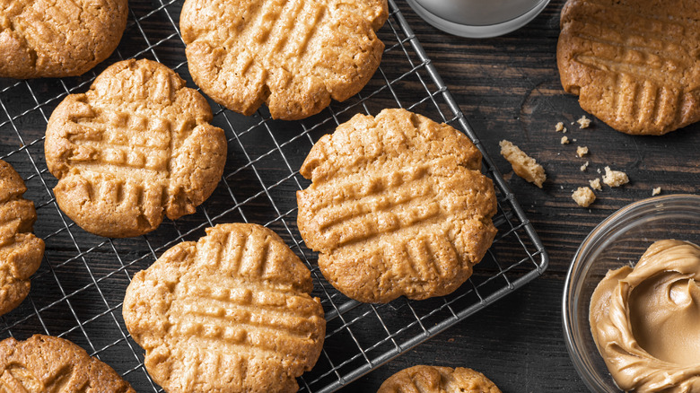 Peanut butter cookies on a wire rack next to a small glass bowl of peanut butter on a dark wood surface