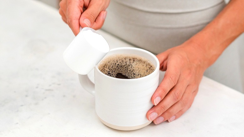 Person adding a scoop of white powder to a cup of black coffee