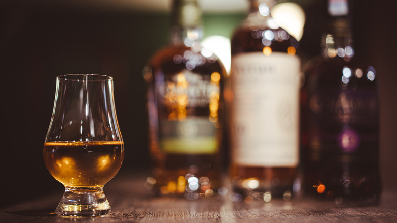 A glass of scotch is photographed with bottles in the background.