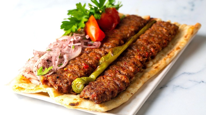 Flat bread with meat kebabs, roasted peppers, chopped tomatoes, and sliced onions on a white plate.