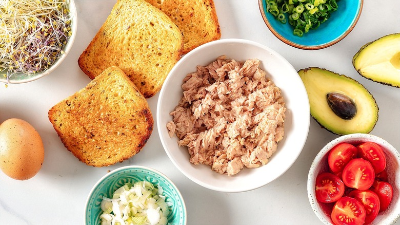 Canned tuna in a white bowl surrounded by pieces of toast, a halved avocado, and small bowls of chopped green onions, sprouts, and halved cherry tomatoes