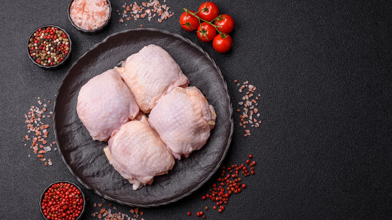 Four raw, skin-on chicken thighs on a black plate surrounded by pink salt, multi-colored peppercorns, and cherry tomatoes on the vine.