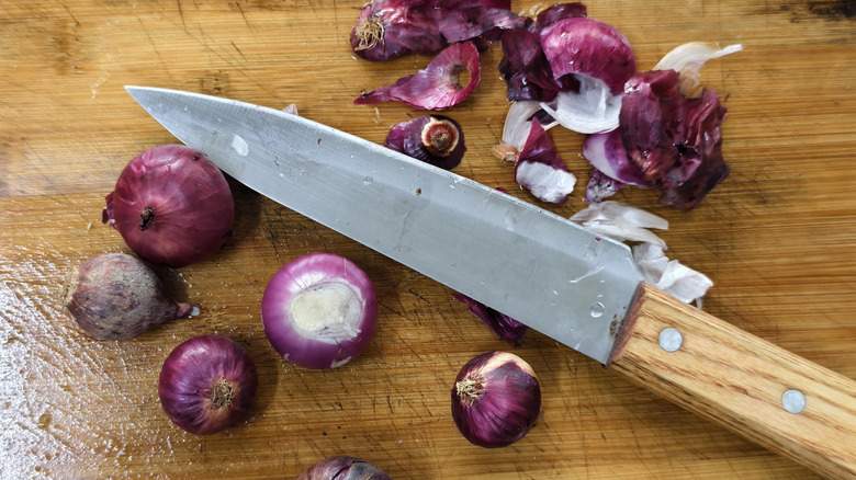 High-angle shot of whole red onions, onion peels, and a chef's knife with a wooden handle on a rustic bamboo
