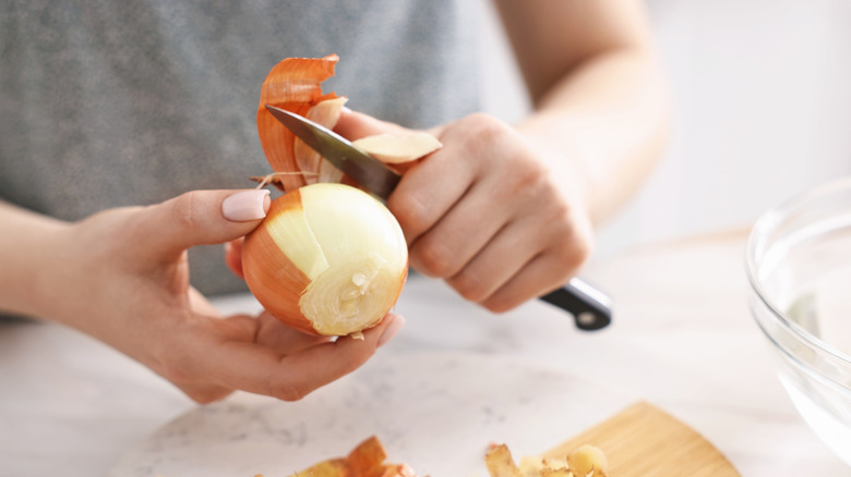Person peeling fresh onion at white table indoors, closeup