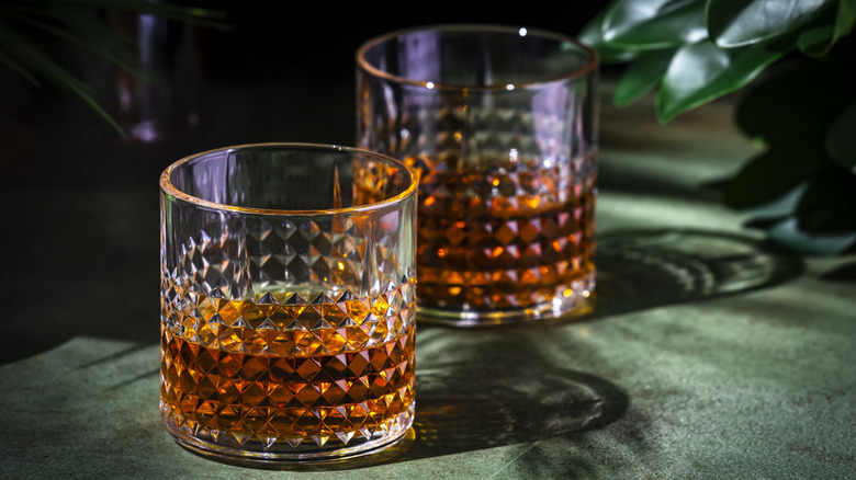 Two rocks glasses of whisky on green counter next to plant.