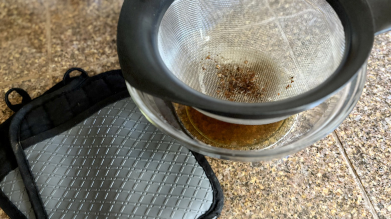 Bacon grease straining through a metal strainer next to a pair of hot pads