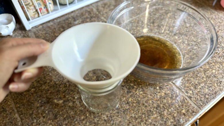 A person holding a wide-mouthed funnel near a glass bowl of bacon grease