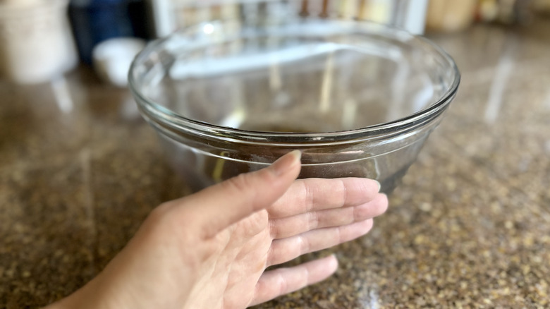 A person's hand testing the temperature of a glass bowl of bacon grease