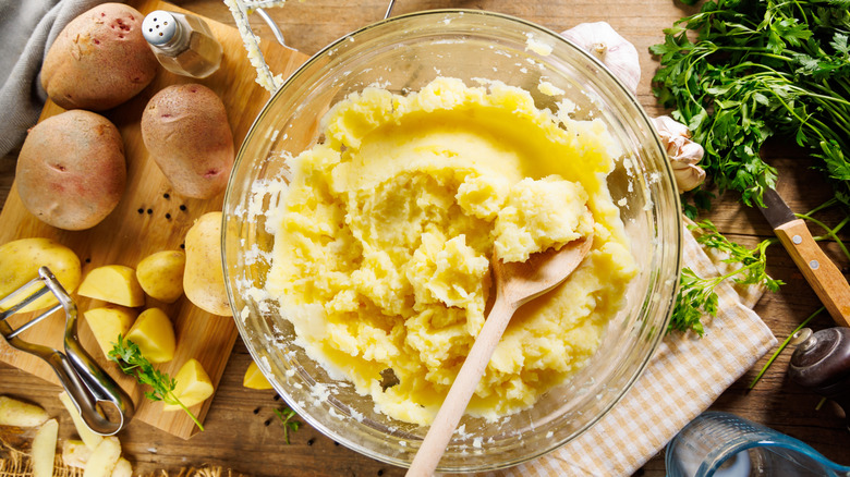 Glass mixing bowl of mashed potatoes with a wooden spoon surrounded by potatoes, parsley, and garlic